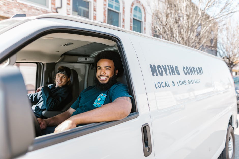 A man with curly dark hair and a beard, dressed in a teal shirt, is standing inside a bright, spacious room with a wooden ceiling and large arched windows. He is positioned behind a stack of large car