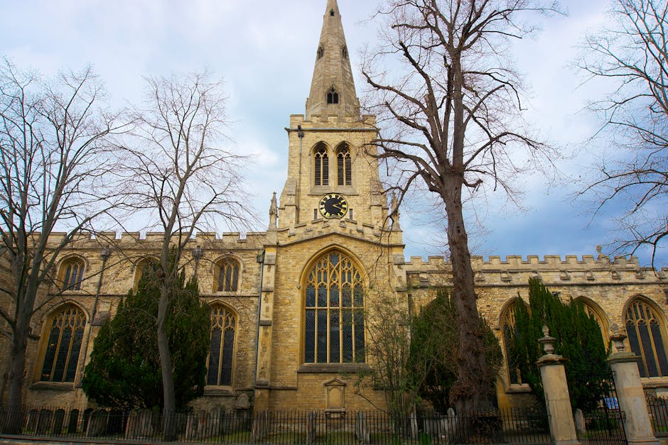 The exterior view of a historic stone church with tall, arched stained glass windows and a prominent clock on the front facade, situated behind leafless trees on a partly cloudy day. The church featur