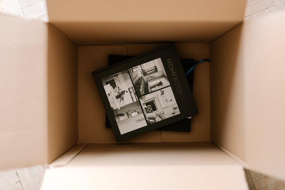 Inside a cardboard moving box, a black photo album or booklet titled 'HOMEBOOK' with black-and-white images of home interiors, including a living room with a sofa, a dining area, and decorative elemen