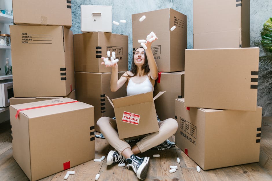 Inside a residential property during a home relocation, a loader wearing protective gloves is seen lifting a cardboard box filled with household items, including a small appliance and plastic-wrapped