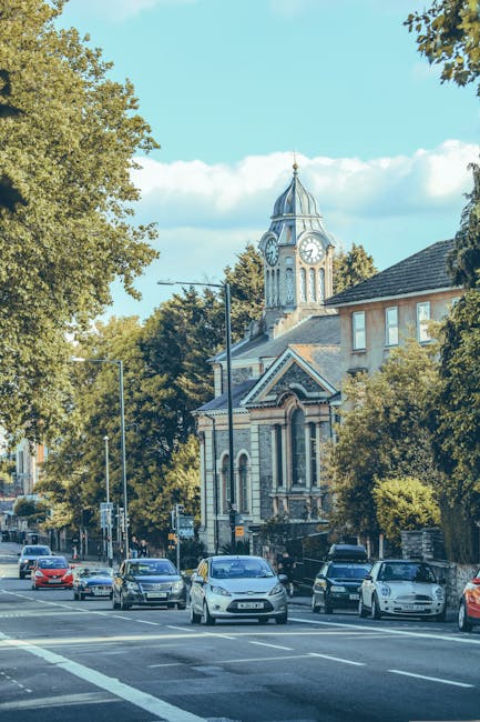 A street scene in Cowley with several parked cars along the roadside and a few moving vehicles on the road, positioned in front of a historic building with a clock tower and a domed roof. Tall trees with lush green foliage line both sides of the street, partially obscuring some of the buildings. The sky is bright and partly cloudy, illuminating the scene with natural daylight. This setting can reflect the neighborhood environment involved in home relocations or furniture transport services offered by Man and Van Cowley. The image captures the urban area suitable for logistics during house removals or moving logistics, showing both the environment and typical street activity relevant to relocation projects and scheduled moving services.