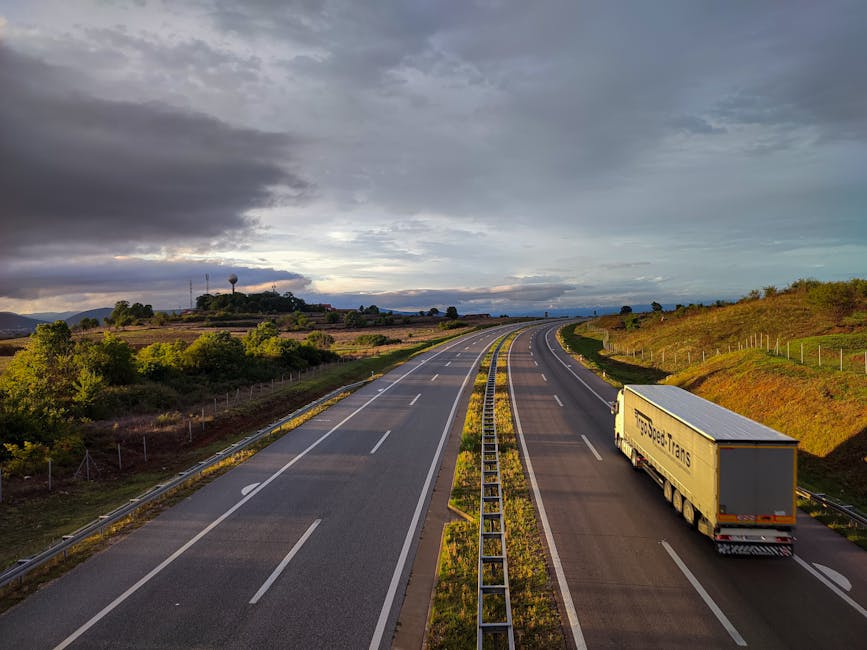 A stretch of four-lane motorway with a central steel crash barrier, taken from an elevated viewpoint. The sky above features a mix of dark, heavy clouds and areas of brighter, clearer sky, indicating changing weather conditions. On the right side of the road, a large white moving truck marked 'Transspeed' is parked in the shoulder, likely involved in home relocation or furniture transport services. The truck is positioned with its back facing away from the camera, ready for loading or unloading. Surrounding the motorway are gently rolling hills covered in grass and sparse shrubbery, with some fencing visible along the roadside. In the background, on the distant horizon, partly obscured by the clouds, are hills or small mountains, along with some trees and man-made structures. The scene depicts a quiet, open stretch of road suitable for long-distance moving logistics managed by companies like Man and Van Cowley within their house removals operations.