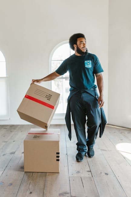 Inside a white moving van with the side door open, two movers are seated in the driver's and passenger's seats. The driver, a man with dark curly hair and a beard, is smiling at the camera, wearing a