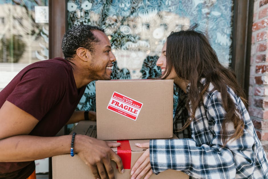 A young woman with long dark hair, dressed in casual clothing, is sitting cross-legged on a wooden floor inside a room, surrounded by numerous large cardboard moving boxes. She is smiling and looking