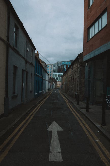 A narrow alleyway between two brick buildings, used as a loading area for house removals. The asphalt ground features double yellow lines along each side, indicating no parking zones. At the end of th